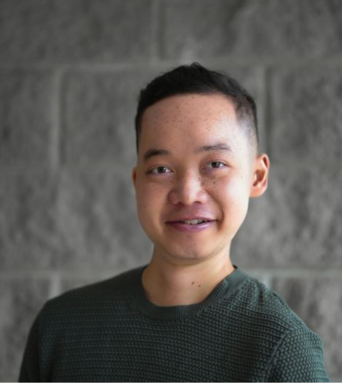 Close up photo of Peter smiling to camera. He wears a green long sleeve and has short dark hair. He stands in front of a bluestone wall.