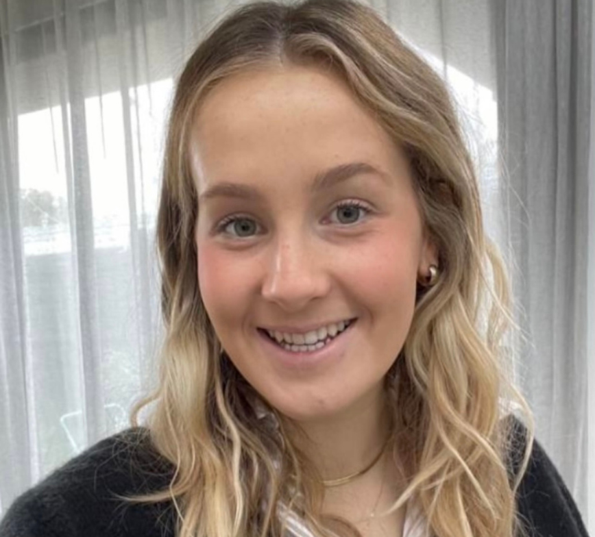 Close up photo of Mia smiling to camera. She has blonde hair and stands in front of a window with sheer curtains.