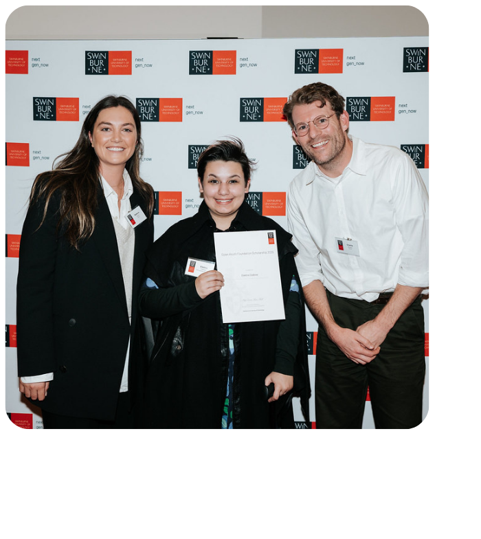 Elektra pictured holding a scholarship certificate in front of a Swinburne Uni media all. She is flanked by DAF staff members Stef (left) and Charlie (right) - all smiling to camera. Elektra holds a walking cane in one hand. 