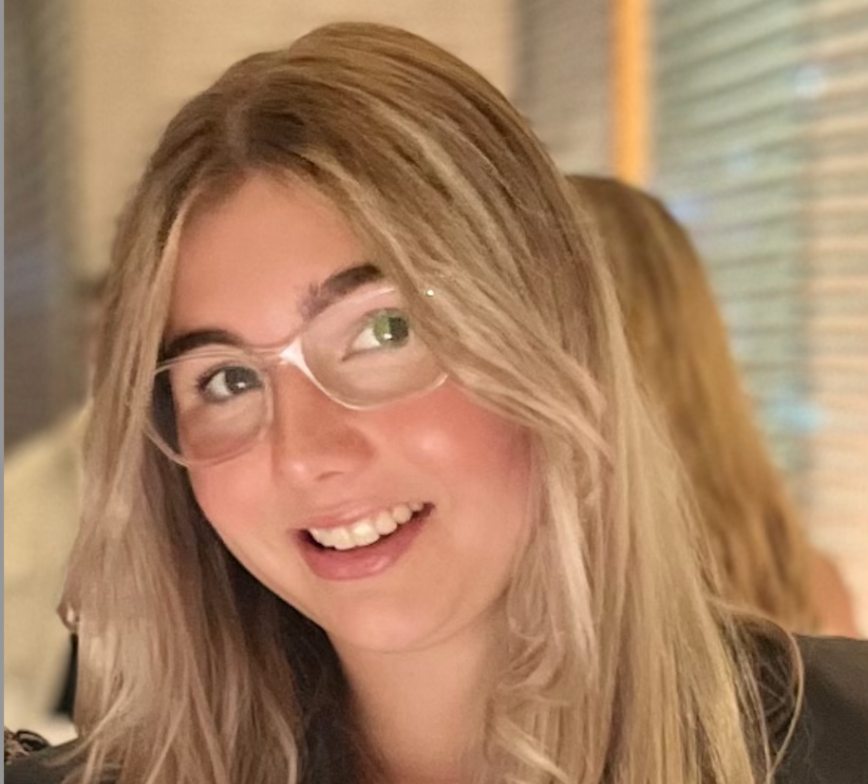 Close up photo of Sienna, she tilts her head to she side and smiles at the camera. She wears clear glasses and the background is blurred.