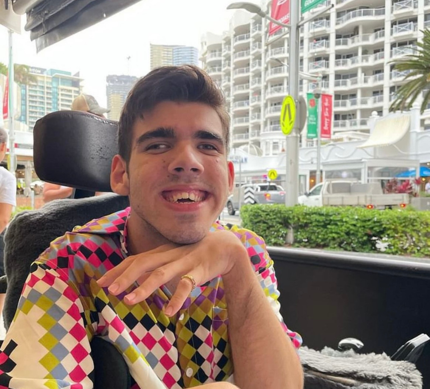 Max uses an electric wheelchair for mobility. He wears a vibrant checkered shirt and smiles to camera with one hand propped under his chin. The background shows white high rise buildings.