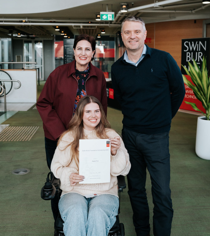 Hayley pictured holding a scholarship certificate in a foyer. She is a wheelchair and parents mum (left) and dad (right) stand behind her smiling to the camera.