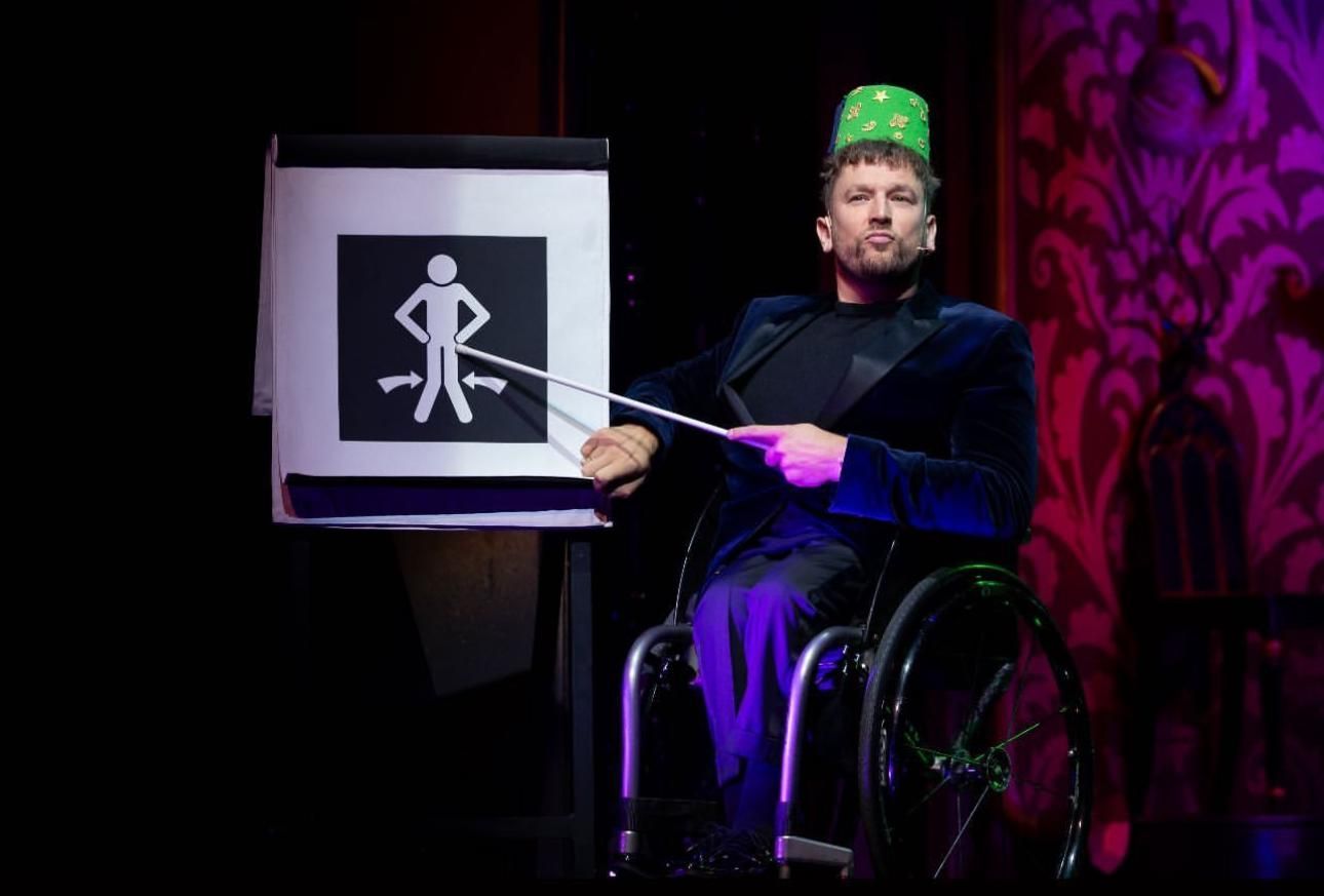 Dylan mid performance on stage using a pointer to point at a diagram on an a frame stand. He wears a green hat and blue velour suit jacket.