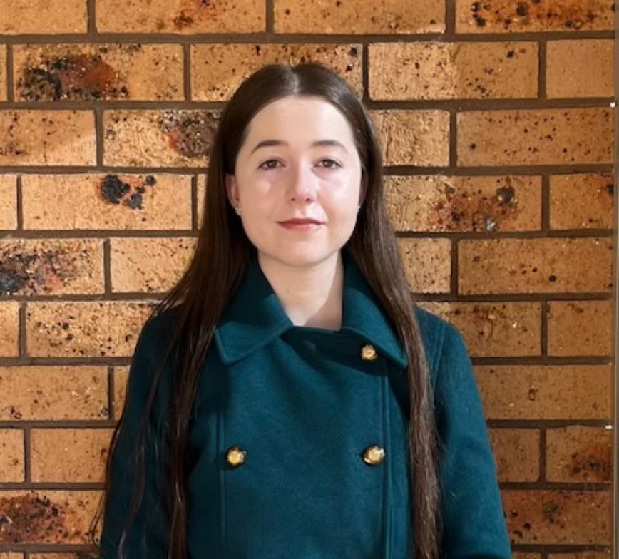 Charlotte stands facing the camera in a green double breasted jacket against a brown brick wall.