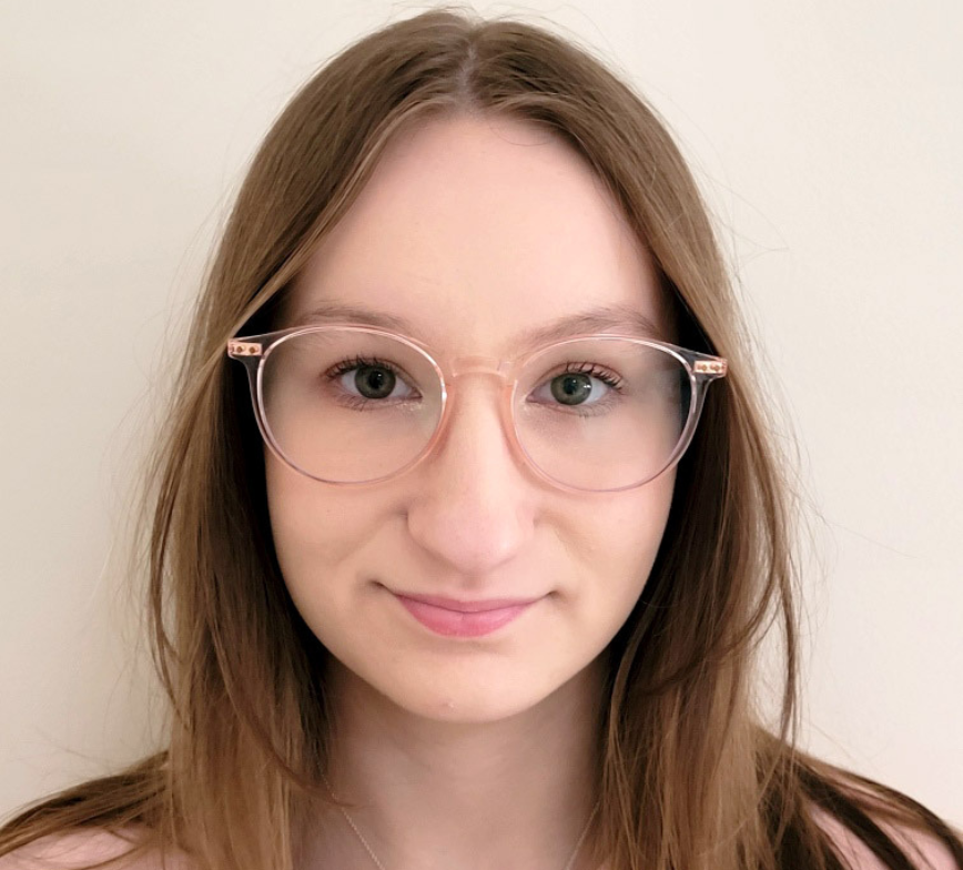 Close up shot of Michala front on smiling to camera. She wears light pink glasses and has light brown hair. The background is a white wall.