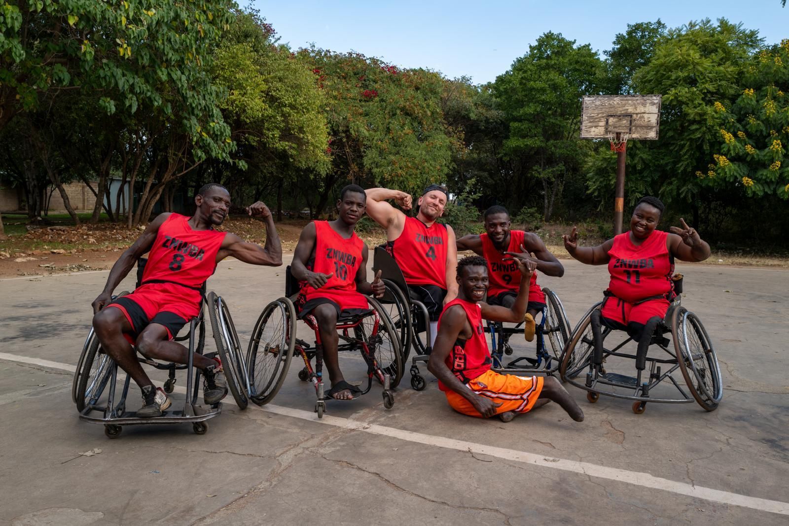 Dylan and a group of young wheelchair basket ballers flex their muscles posing for the photo. They all wear red basketball jerseys and are on an outdoor basketball court.