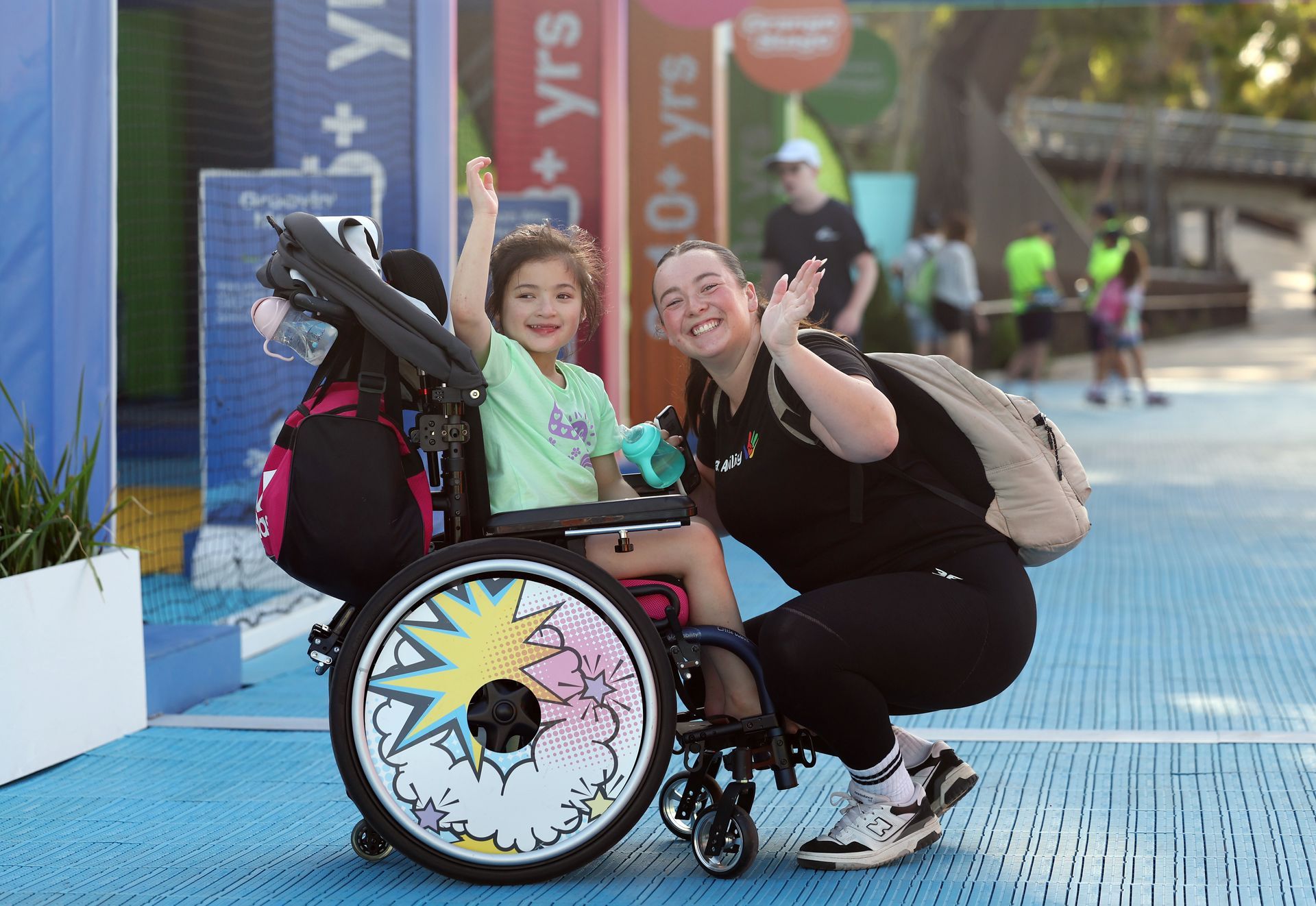 A young girl who is a wheelchair user and a woman who crouches down in font of her. Both are turned to the side smiling and waving at the camera. The background shows the AO Ball Park.