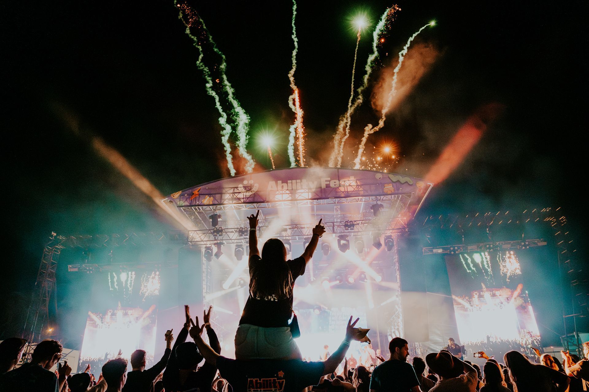 Night time photo of a female sitting on a mans shoulders facing the Ability Fest main stage. Both their hands are thrown in the air in celebration. The background shows the stage lit up and fireworks streaming from the top.