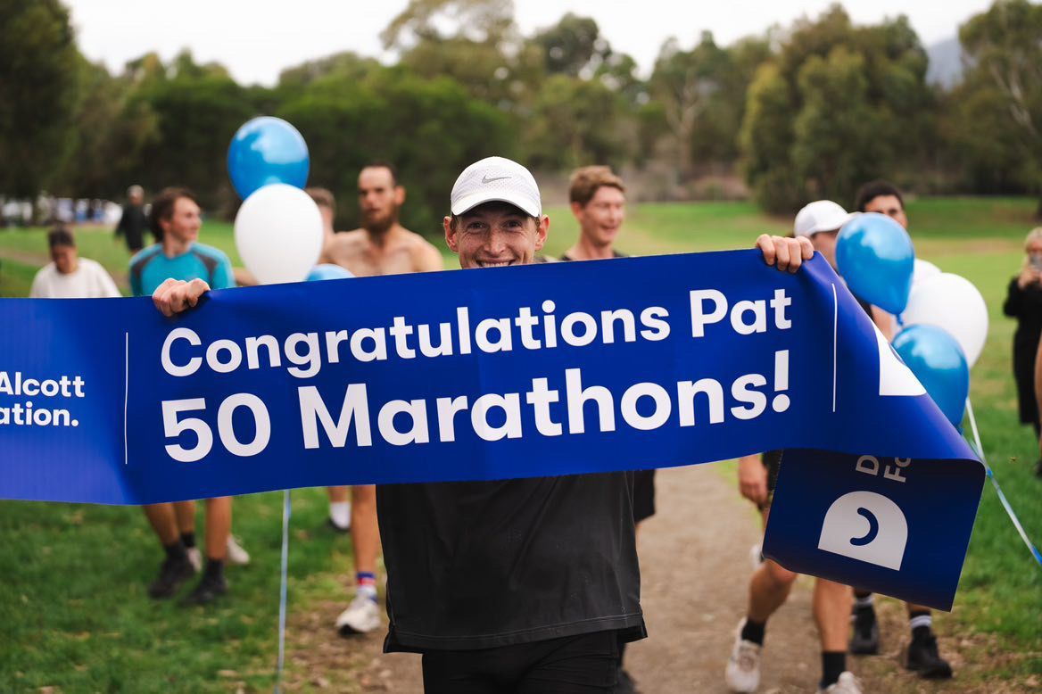 Pat runs through a finish line sign that is dark blue and reads - Congratulations Pat, 50 Marathons!