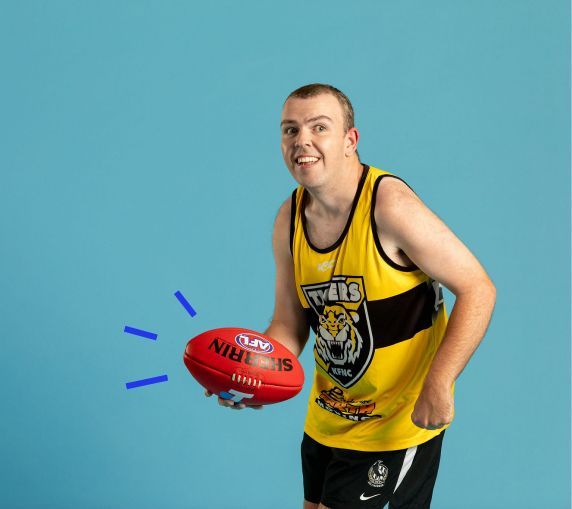 Male wearing a yellow tigers jersey, holding a red sherrin football. He is mid action handball. 