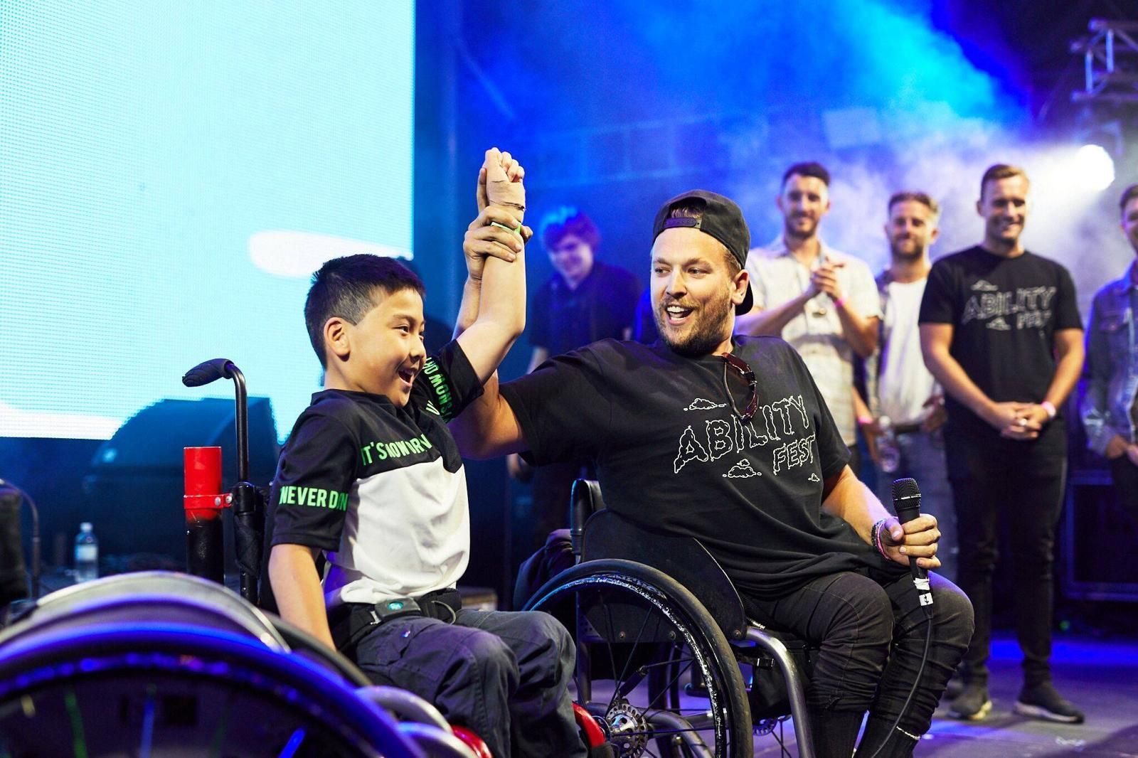 Recipient Jin (left) and Dylan (right) holding hands raised on stage at Ability Fest. Both are wheelchair users. 