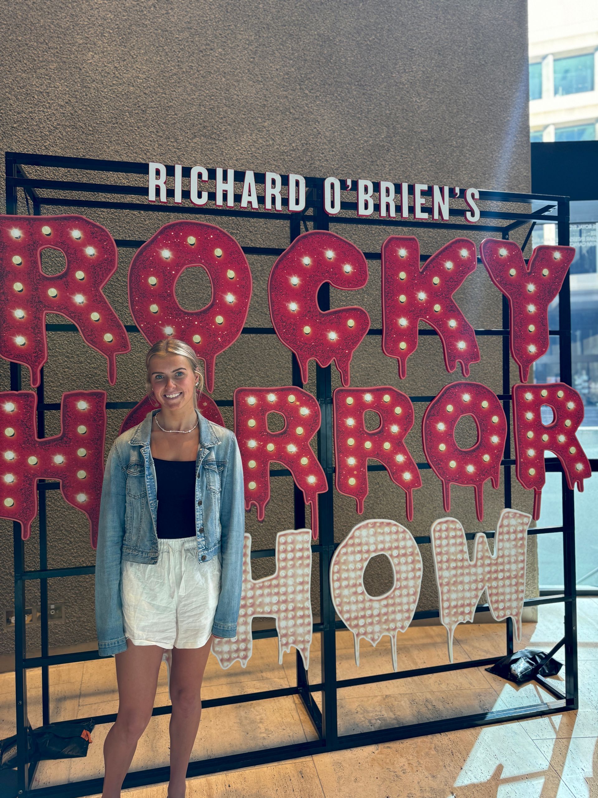Hannah poses for the camera in front of the red and white Rocky Horror sign that reads - Richard O'Brien's Rocky Horror Show.