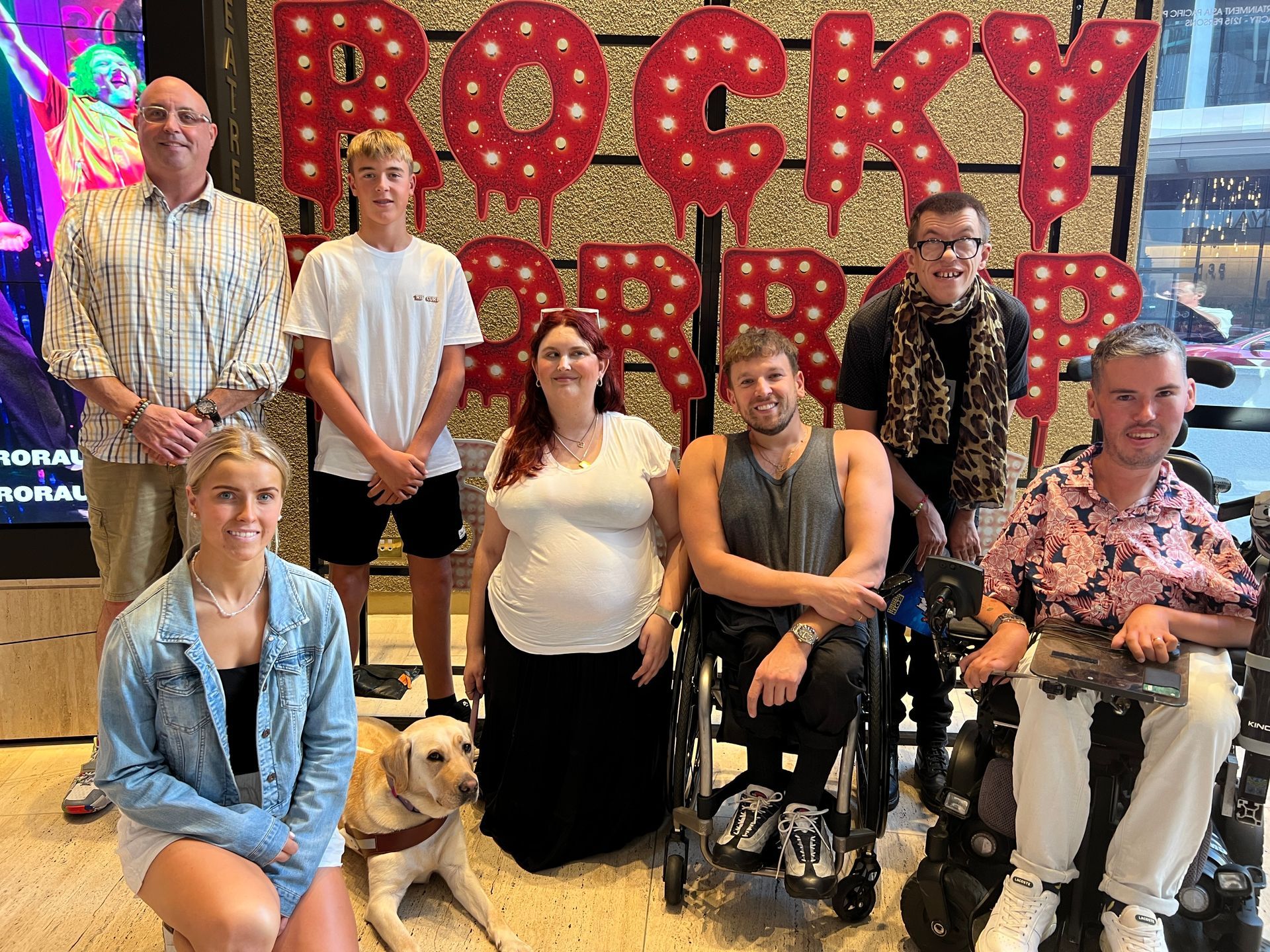 Dylan (middle) poses for a group photo with DAF recipients of varying disability in front of the red Rocky Horror sign that reads - Rocky Horror Show.
