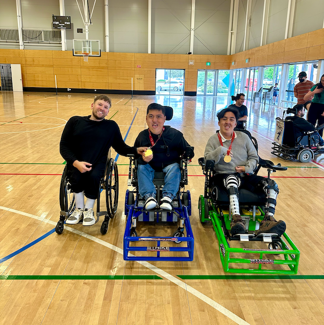 Dylan (left) with recipient Jono (middle) and Josh (right) whom are in strikeforce power chairs. They wear gold medals whilst on an indoor basketball court.