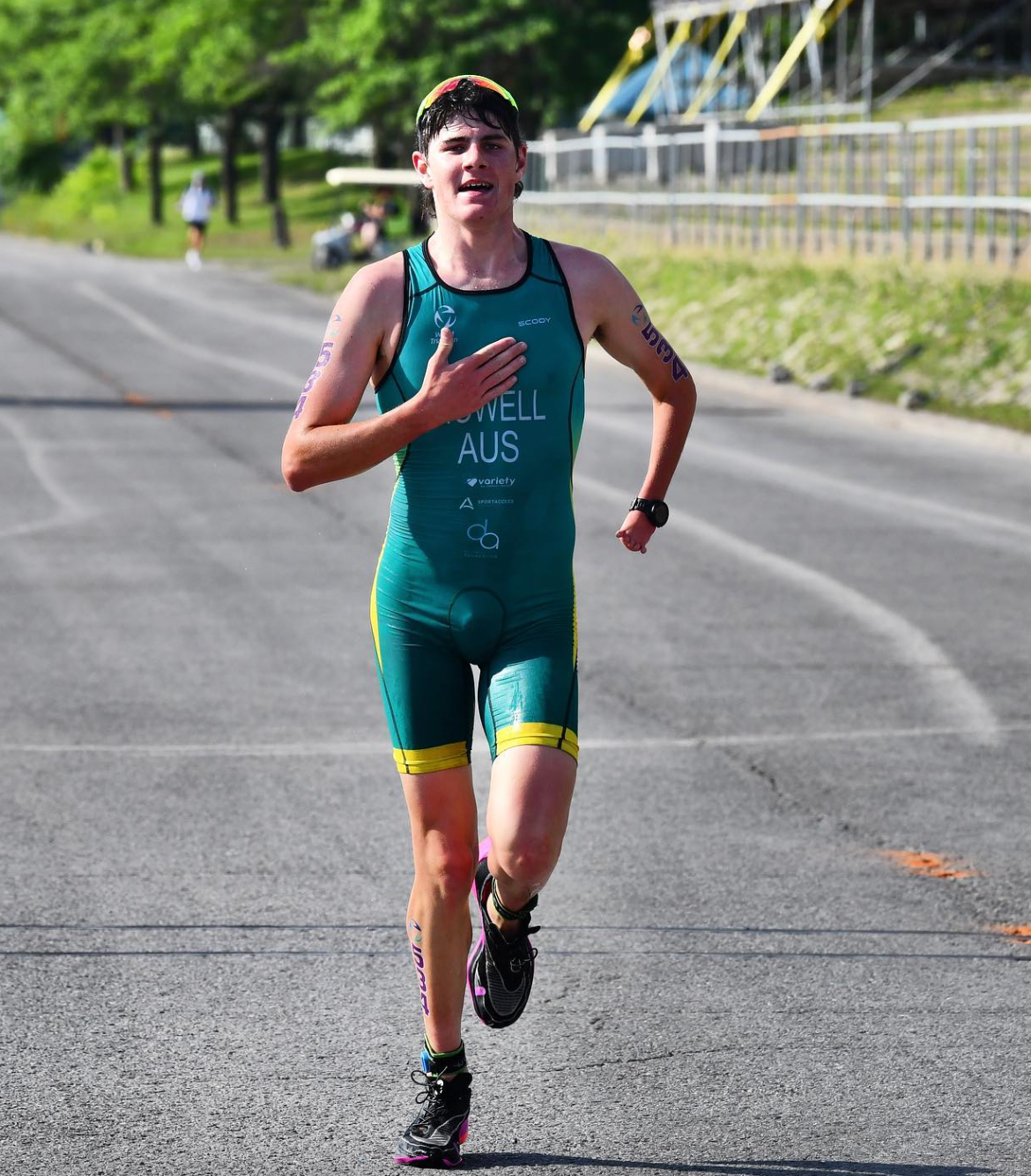 Jack competing in his Australian green and yellow uniform in the running leg of the race. He runs front on toward the camera on a concrete road, wearing black runners, sunglasses a top his head and a black watch on the left wrist. His right hand is placed flat against the chest. The front of his uniform in white writing reads as HOWELL, AUS, followed by sponsors including the DAF logo.
