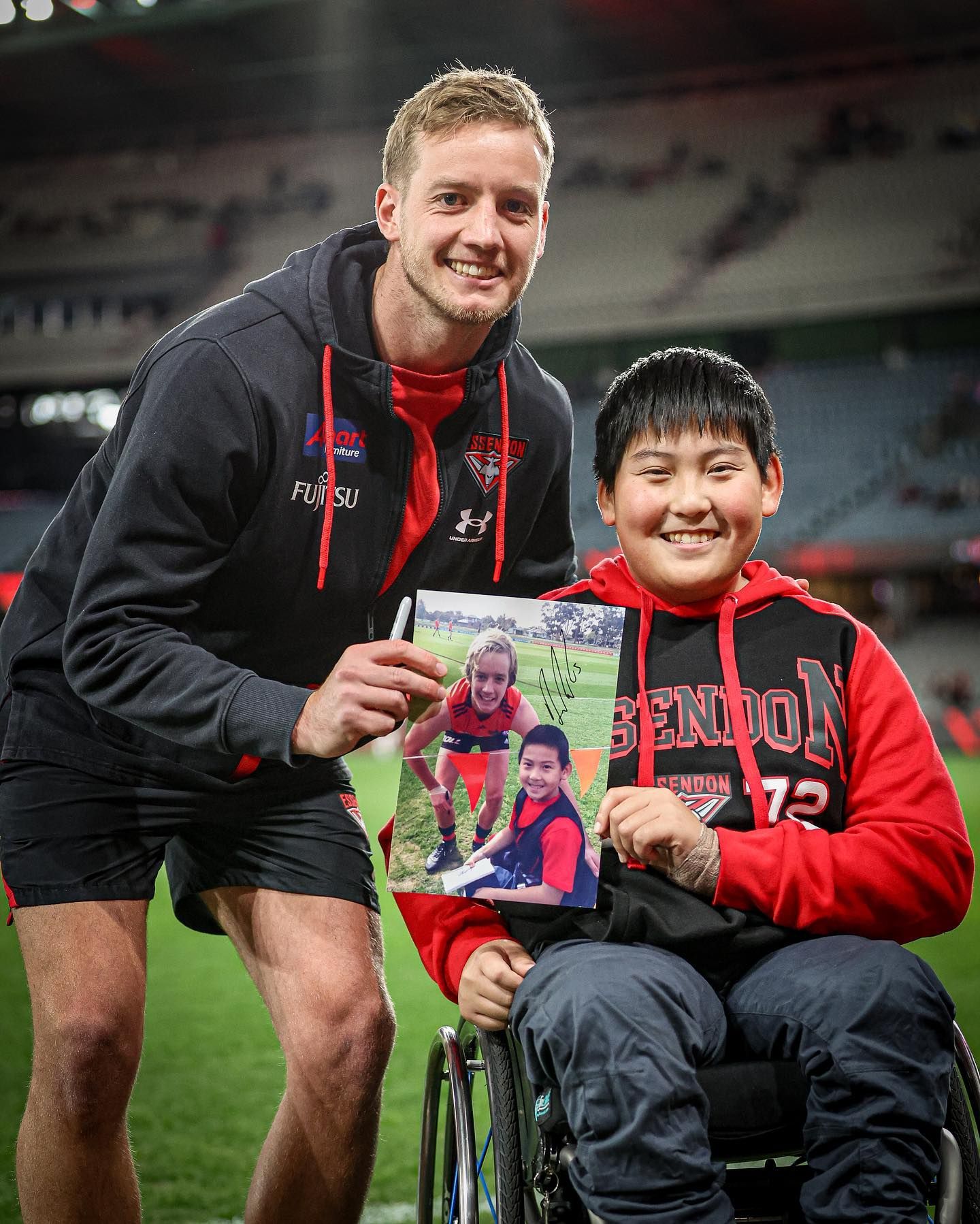 Darcy Parish (left) and Jin Woodman (right) smile to camera on the boundary line at Marvel Stadium. They hold a picture of themselves taken 7 years prior, signed by Darcy.