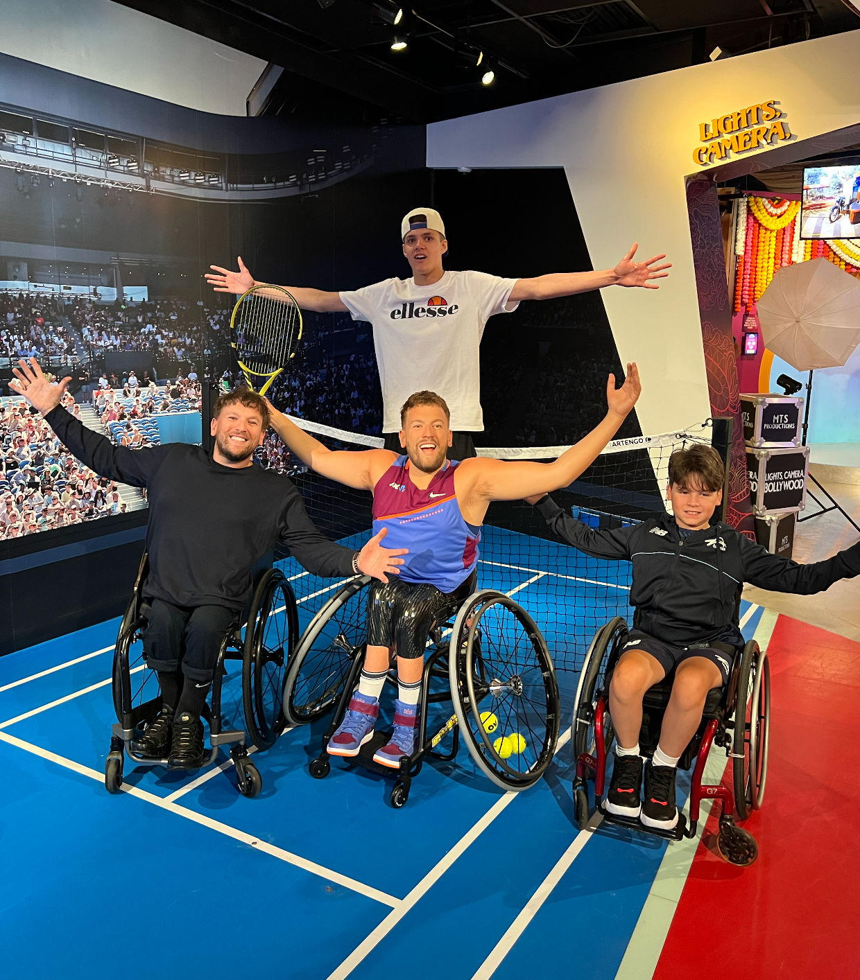 Located on a miniature tennis court - blue ground, white lines and net through the middle. Dylan's wax figurine is dressed in tennis grand slam kit, arms splayed wide, holding his racquet high, smiling. Real Dylan is positioned to the left, with grant recipient Lachlan standing behind and Rafferty to the right in his wheel chair. All three have their hands wide, mimicking the wax figurine.