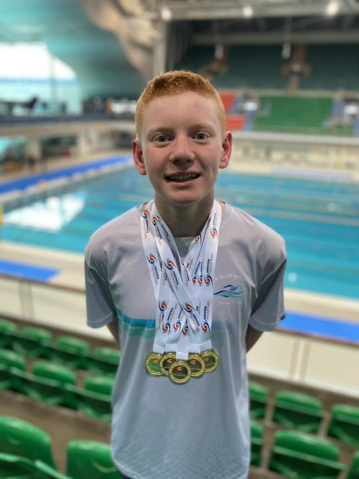 Recipient Noah stands in the grand stand at a swimming centre with multiple medals around his neck.
