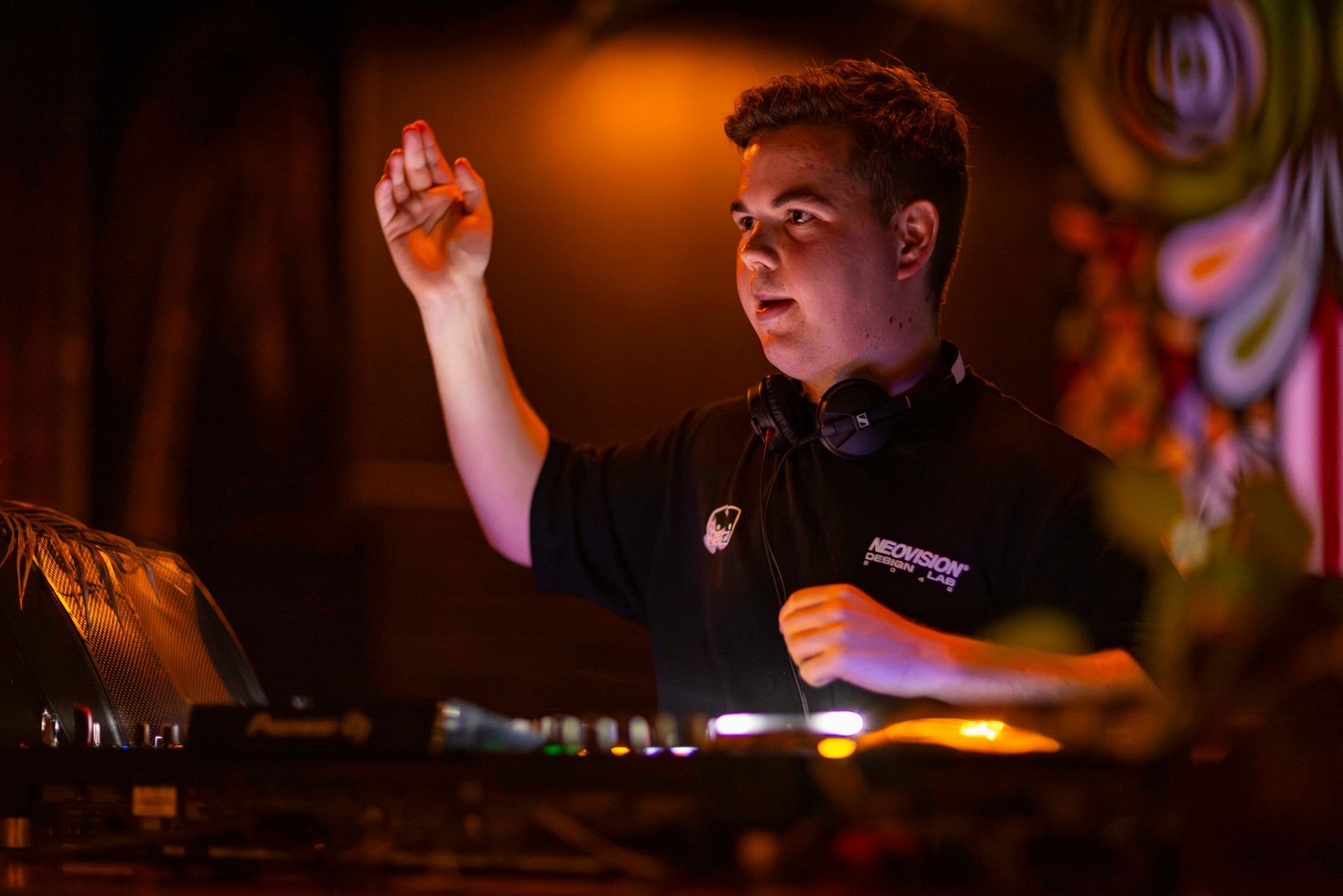 Close up image of Noah standing behind a set of dj decks as he faces the crowd, with one arm raised. He wears headphones around his neck