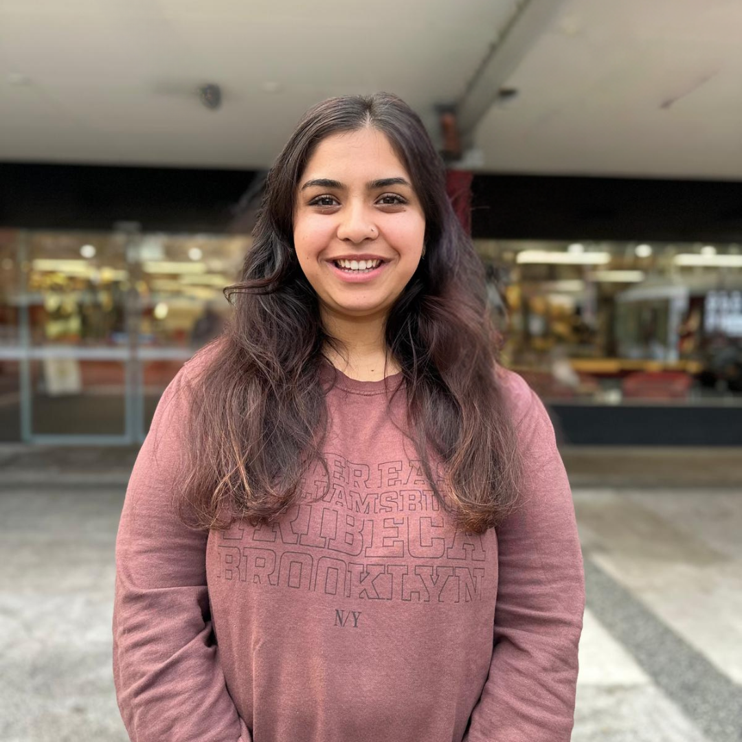 Samiksha smiles to camera out the front of the library university which appears blurred. She wears a burgundy jumper.