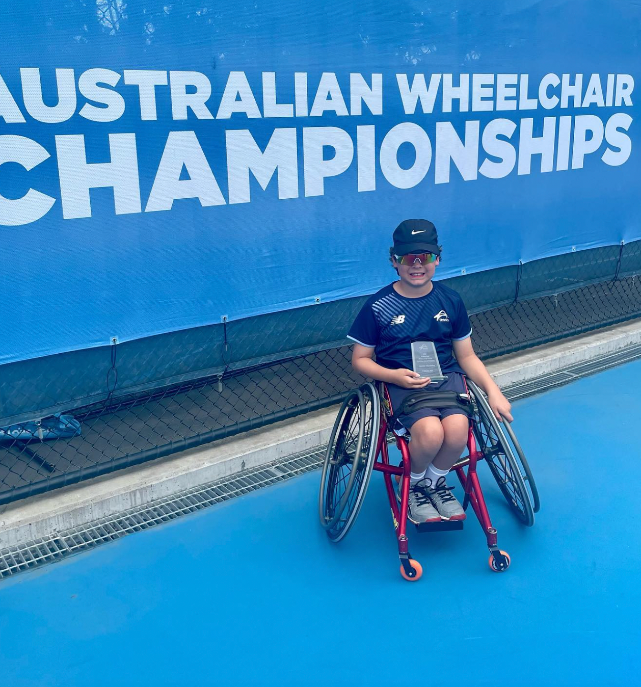 Raf smiles to camera holding a trophy on a blue tennis court. He uses his foundation tennis chair. The word 'championships' is behind Raf on a blue background with bold white letters.