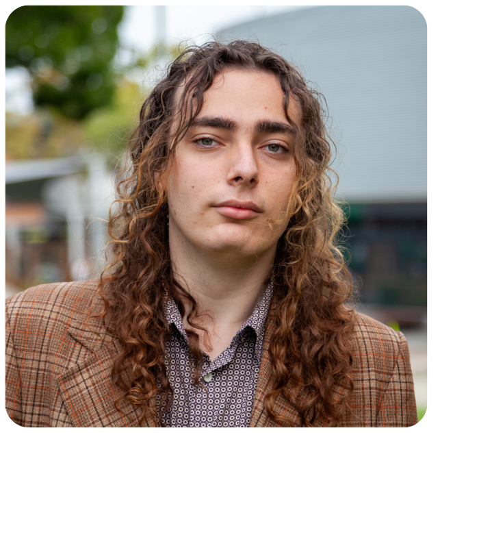 Pictured outdoors with a blurred background, Saul has long brown wavy hair and wears a brown tweed check jacket.