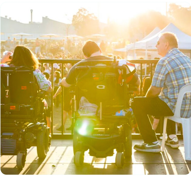 Shot from behind of 3 individuals sitting on the viewing platform at Ability Fest. Two individuals on the left are wheelchair users and Ability Fest crowd in the background.