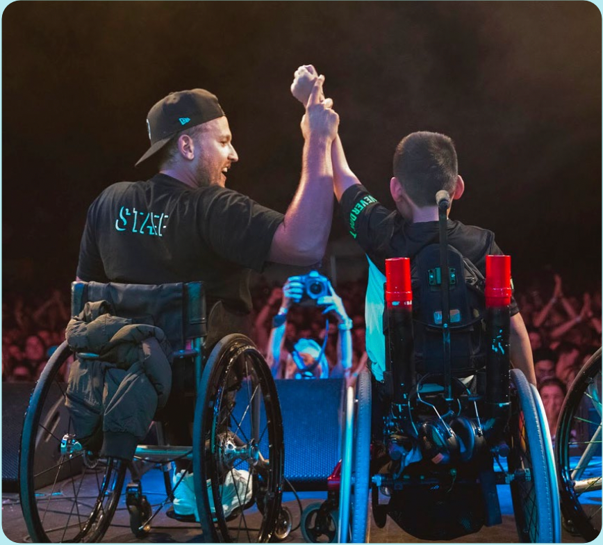 Shot from behind of recipient Jin (right) and Dylan (left) holding hands raised on stage at Ability Fest. Both are wheelchair users. 