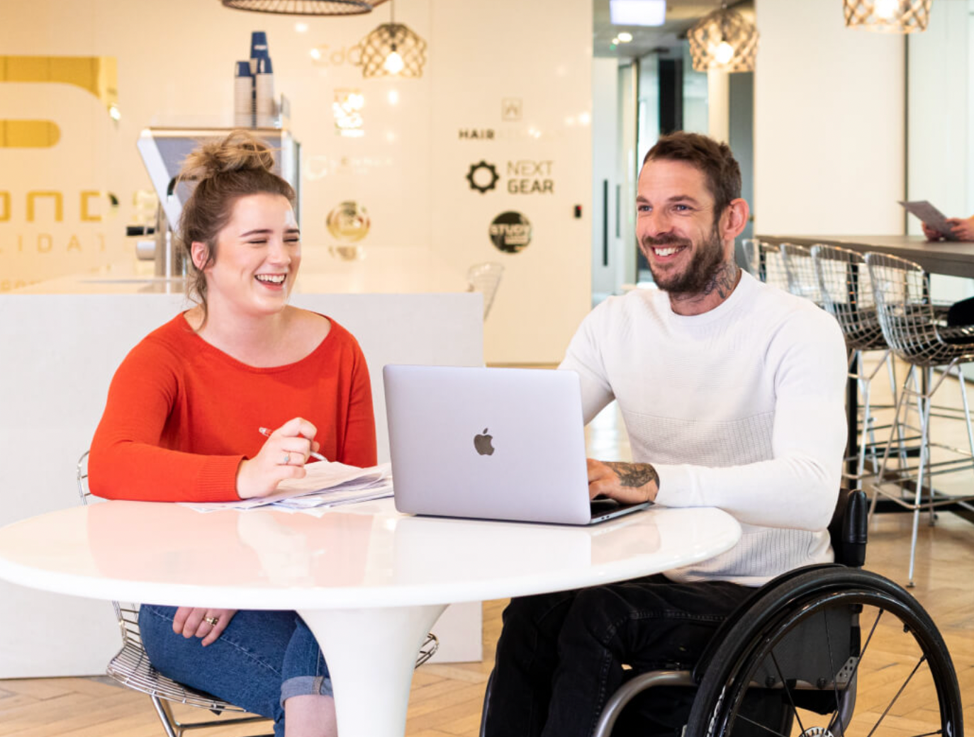 Female (left) sitting at a white office work desk alongside a male wheelchair user (right) who works on his laptop.