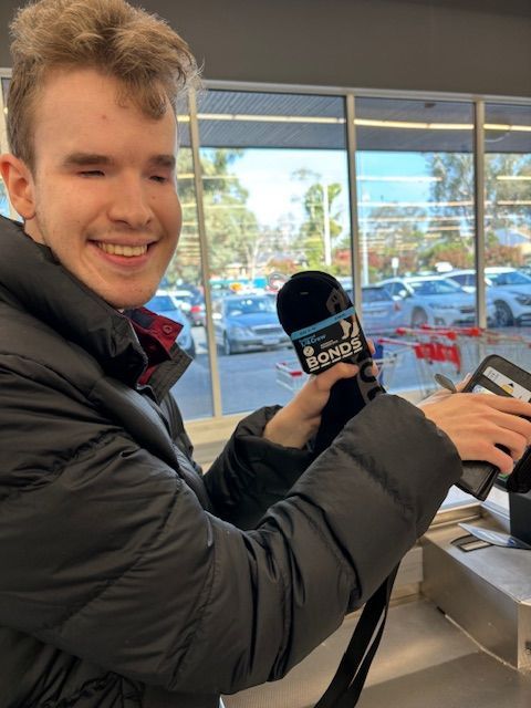 Recipient Riley, poses for photo mid check out holding his Bonds socks in his left hand. He smiles to camera whilst using his phone (right hand) to pay-pass on an eftpos terminal. The background shows a parking lot behind floor to ceiling windows.