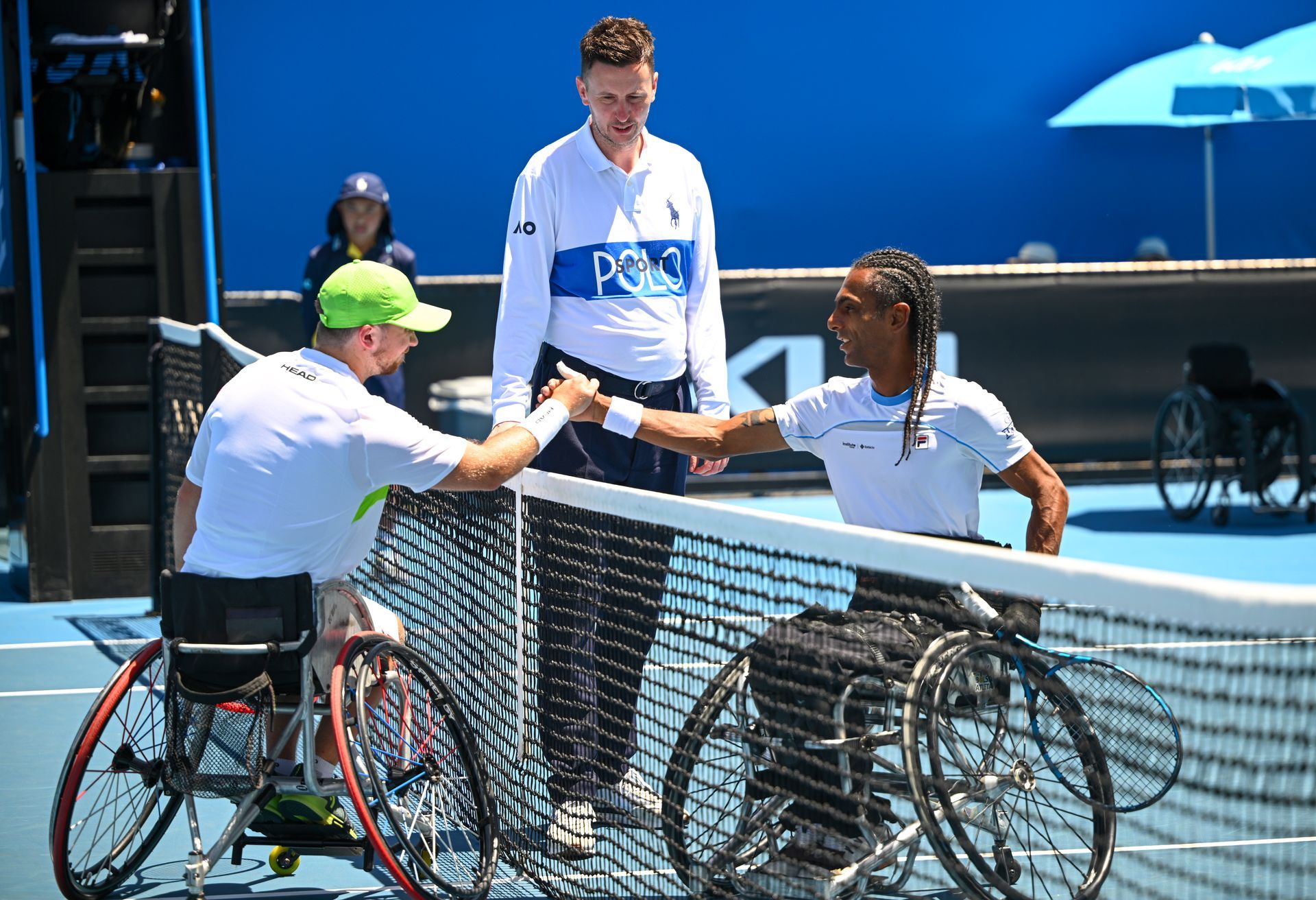 Two male tennis players who are in tennis wheelchairs. They are shaking hands over the net with the umpire standing behind them.