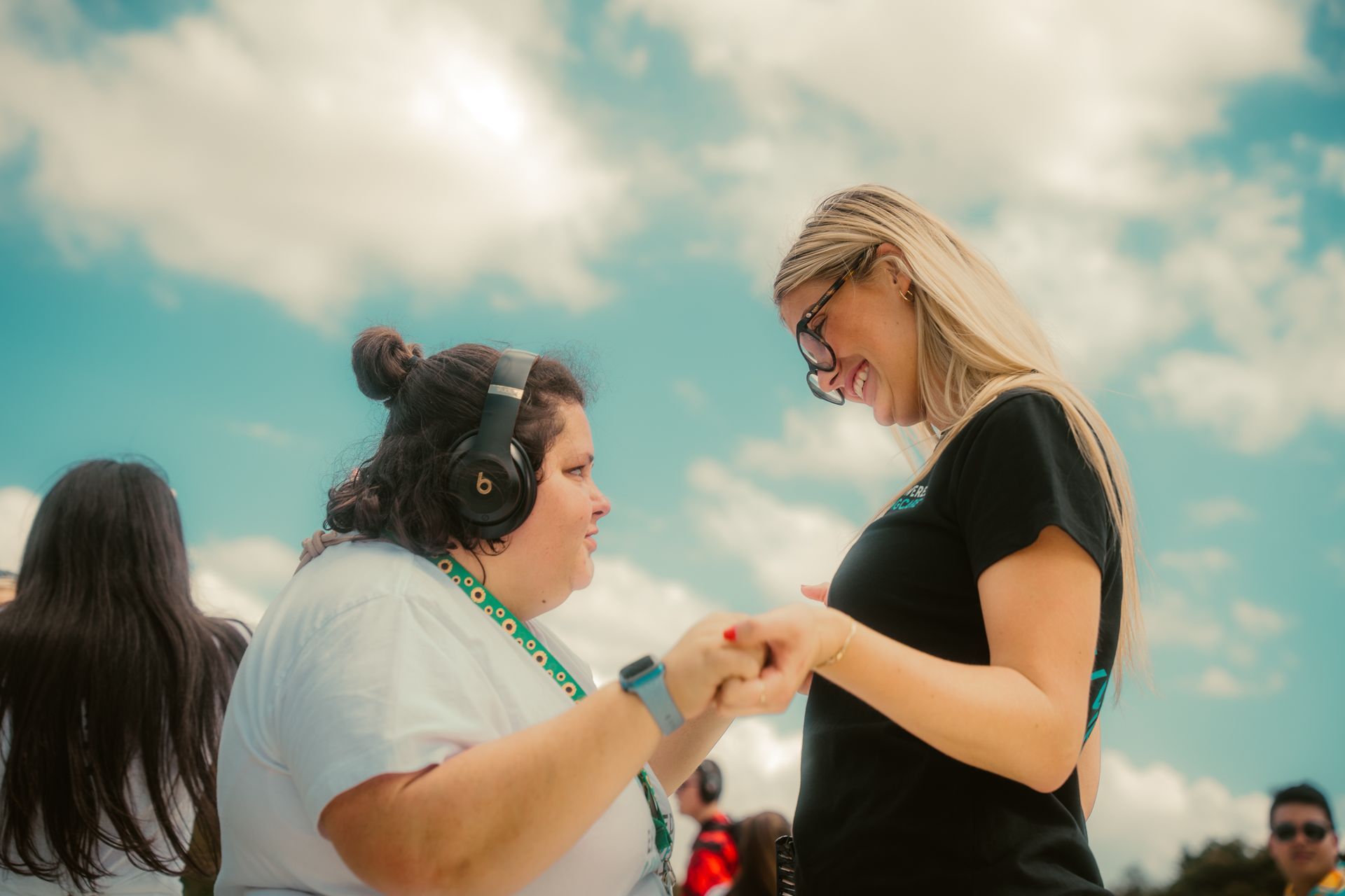 Photo taken outdoors of two women holding hands and looking at each other smiling. One women on the left wears a white t-shirt, a sunflower initiatives lanyard with headphones and the woman on the right wears a black t-shirt and black framed glasses.