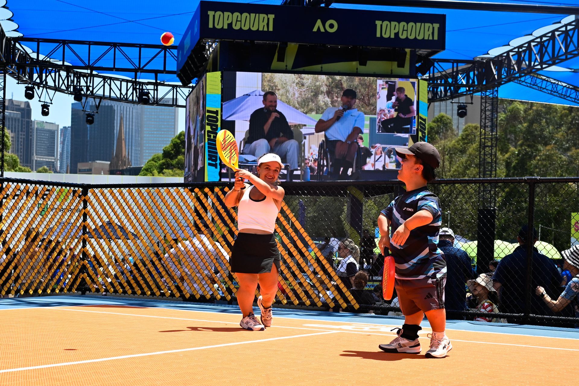 One male and one female playing on a paddle court playing pickle ball. The background shows Dylan and Zack Alcott on a big screen as they conduct an on stage Q&A.