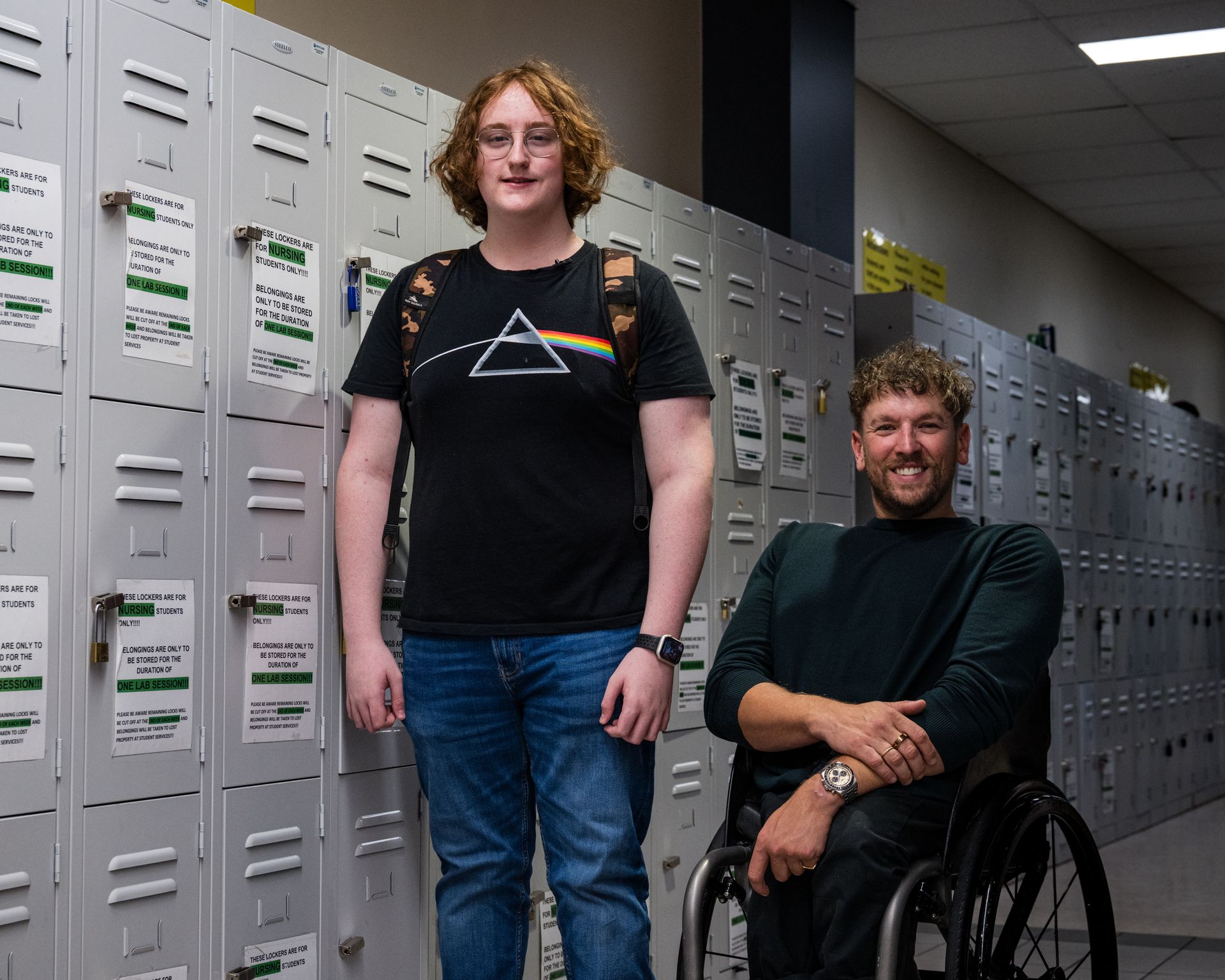 Connor (left) stands side by side with Dylan (right). They smile to the camera in a hallway filled with grey lockers behind.