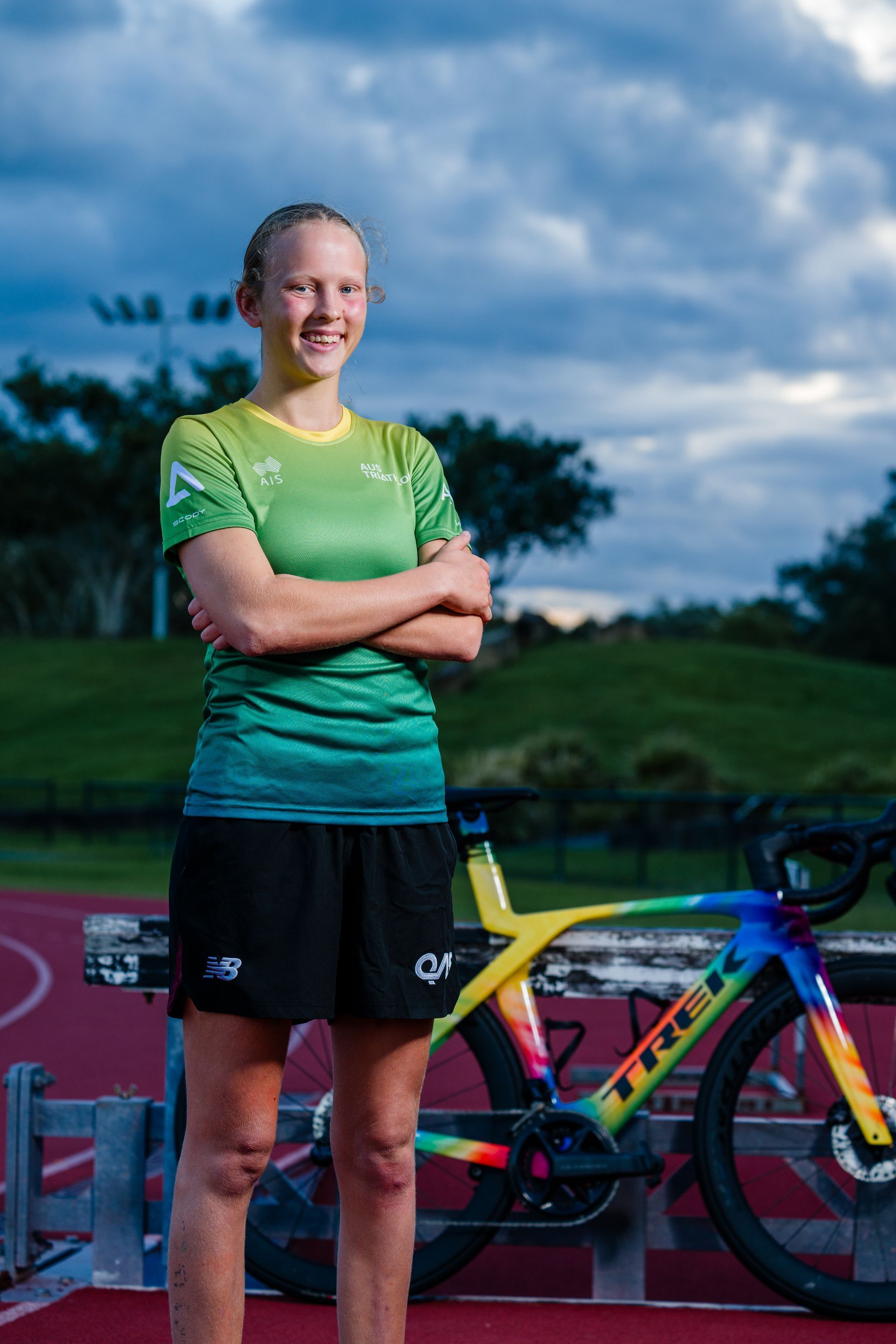 Recipient Grace stands on an athletics track with her arms crossed smiling to camera. Her rainbow bike is resting in the background.
