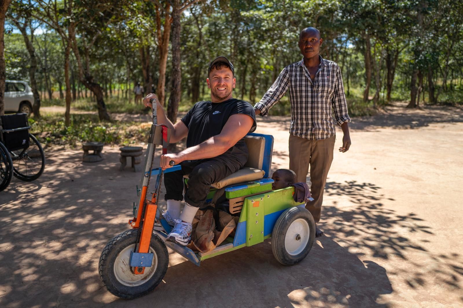 Dylan trialling a home made colourful mobility aid created in Zambia that has hand pedals. A man behind Dylan poses for the photo smiling with trees in the background.
