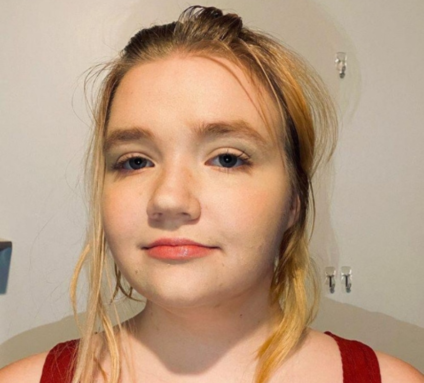 Close up head shot of Daphne. She wears a red singlet top and hear blonde hair up in a bun. She is in front of a white wall.