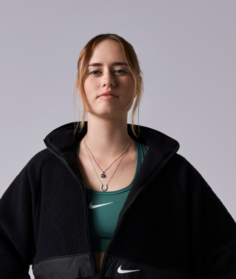 Studio portrait of Georgia, she wears a woolen Nike jumper with her arms on her hips. A green Nike sports bra can be seen underneath.