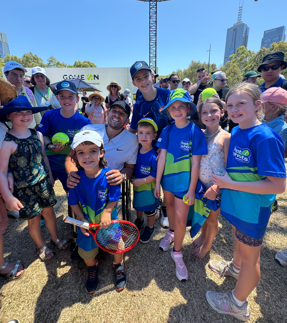 Dylan surrounded by kids wearing blue Tennis Australia t-shirts at the AO Ball Park. They all smile at the camera.