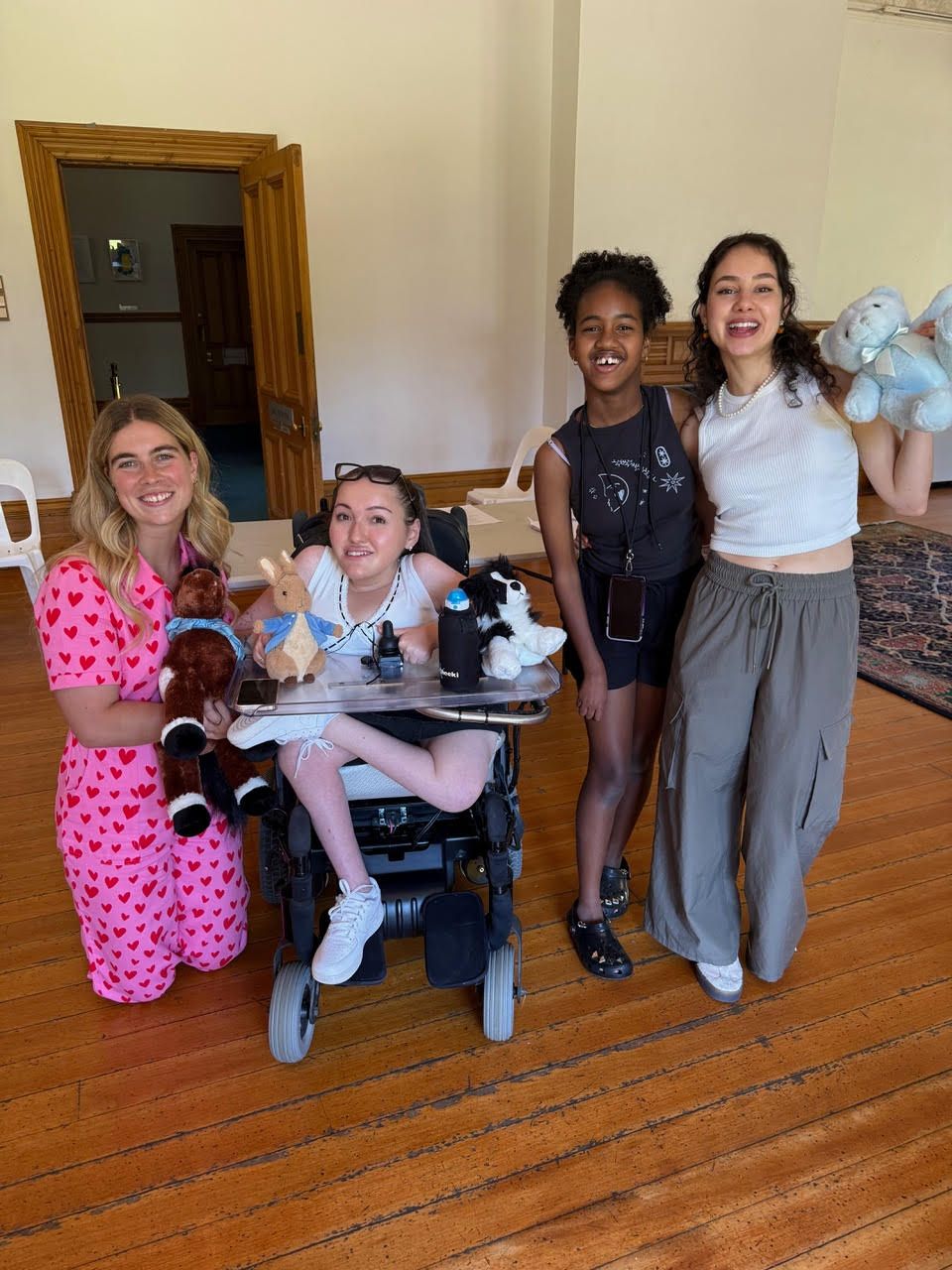 Lily (left) with three female cast members (right) smile to the camera on set. Lily is kneeling down next to a female character who is a wheelchair user, whilst the other two females remain standing in costume.