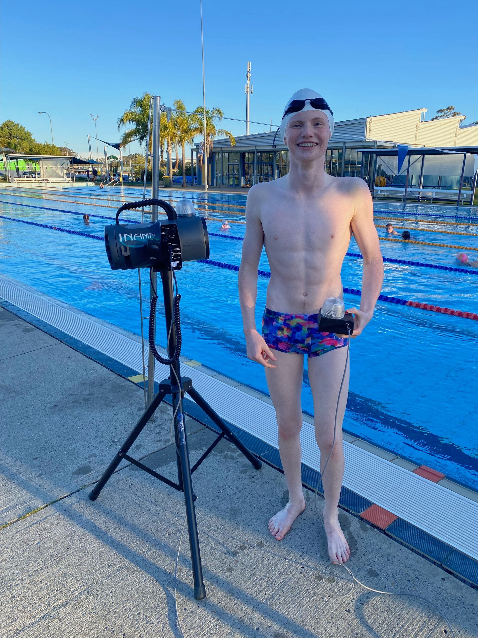 Seb stands at a pools edge in his swimming gear next to the electronic swim start system. He holds the systems connected light.