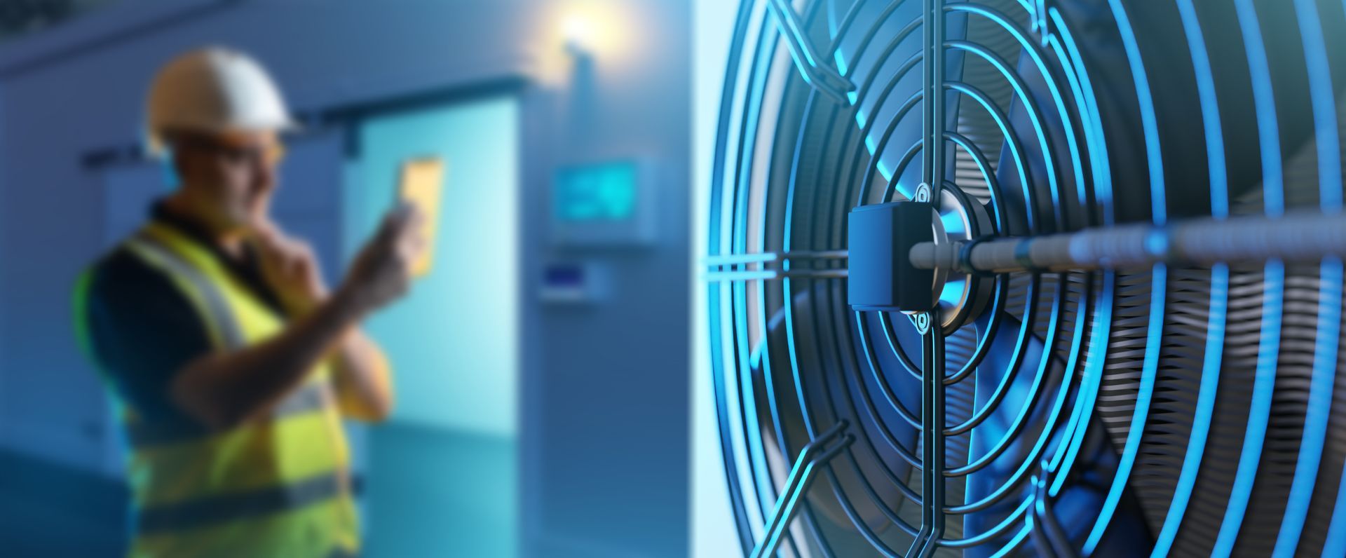 Blurred image of a construction worker in a safety vest holding a phone near a large fan in a mechanical room.