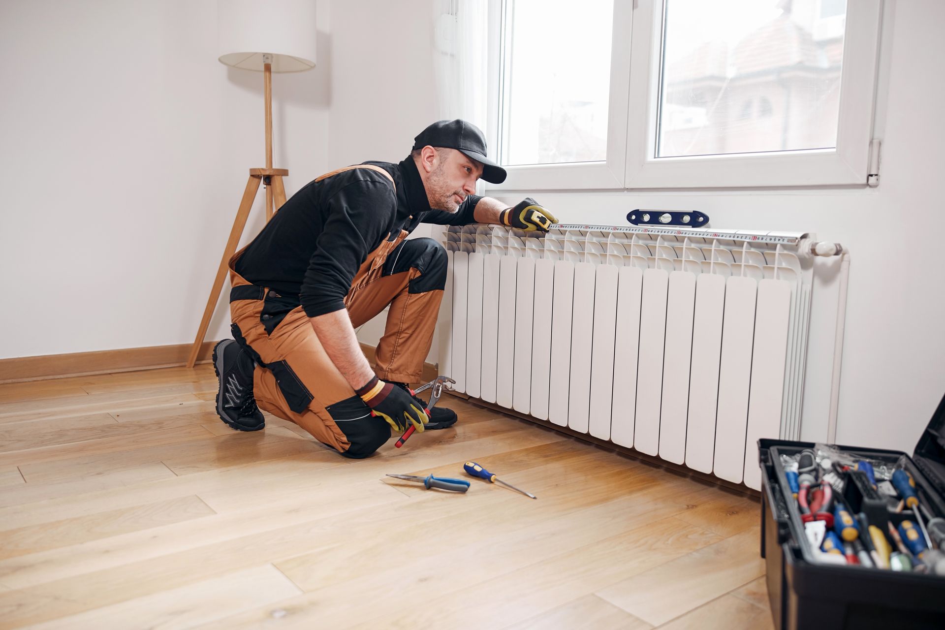 Person kneels by radiator, using tools. Inside a room.