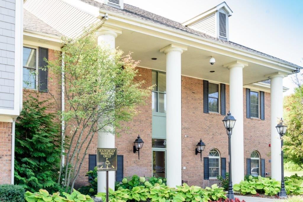 Brick building with white columns and dark shutters, surrounded by green shrubs.