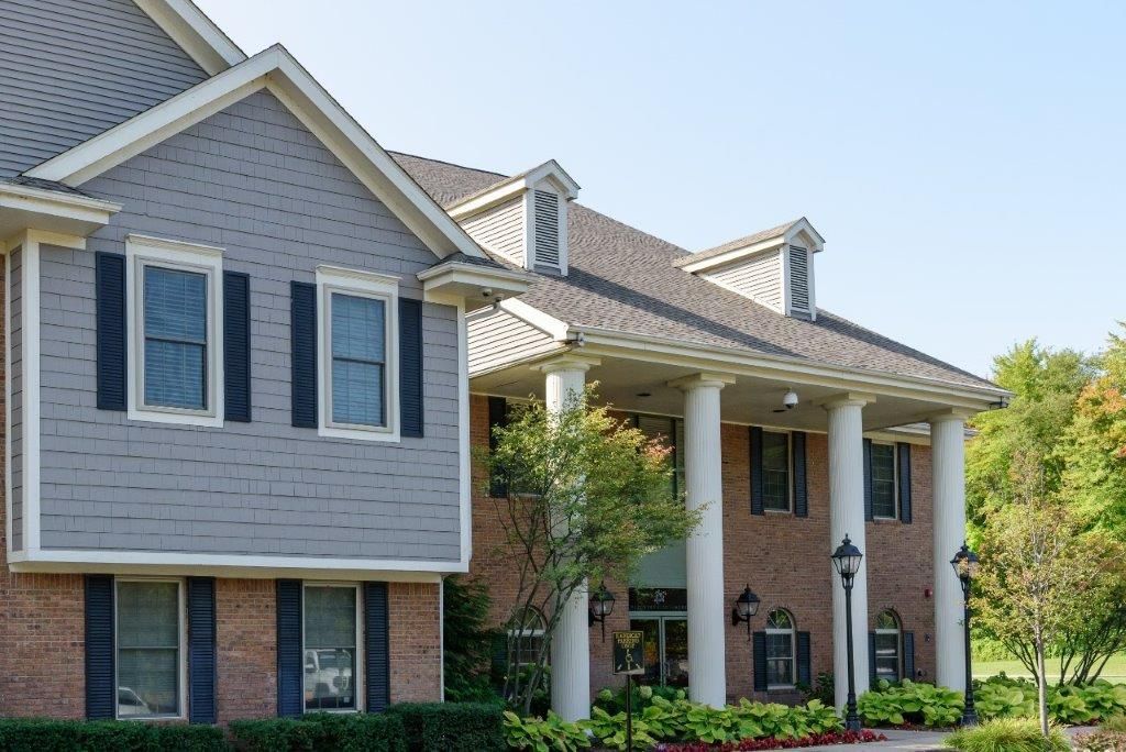 Two-story house with gray siding and brick. Features large white columns, dark shutters, and a sloped roof.