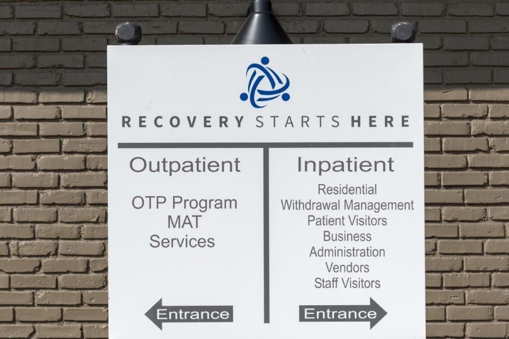 Sign indicating outpatient and inpatient services at a recovery center. Black text on white background with a brick wall.