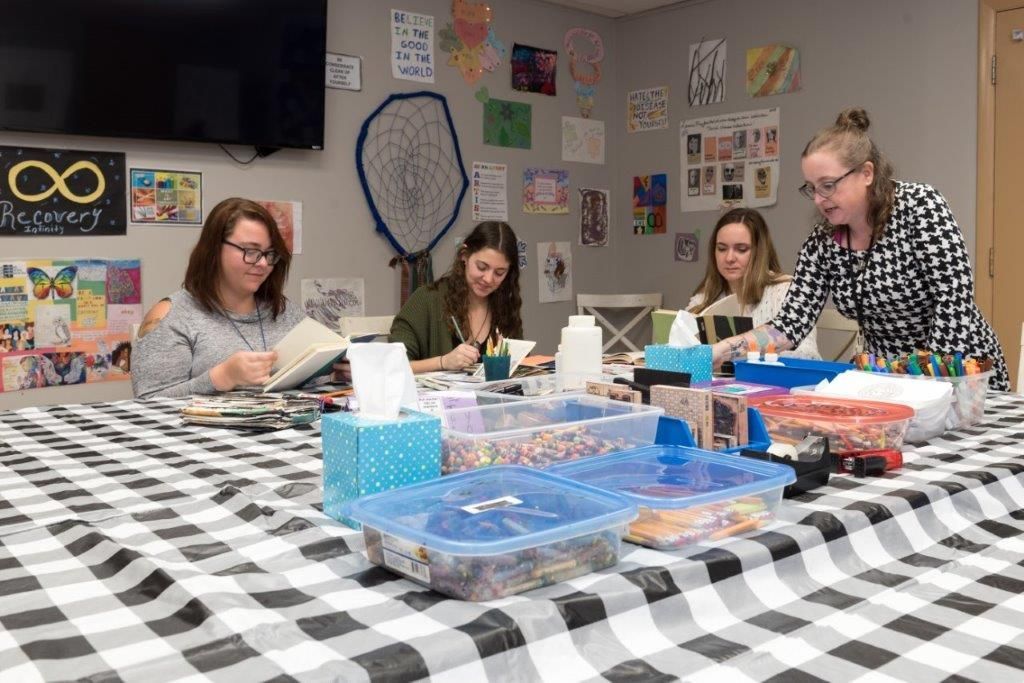 Four people craft at a table with art supplies.  The room has artwork on the walls.