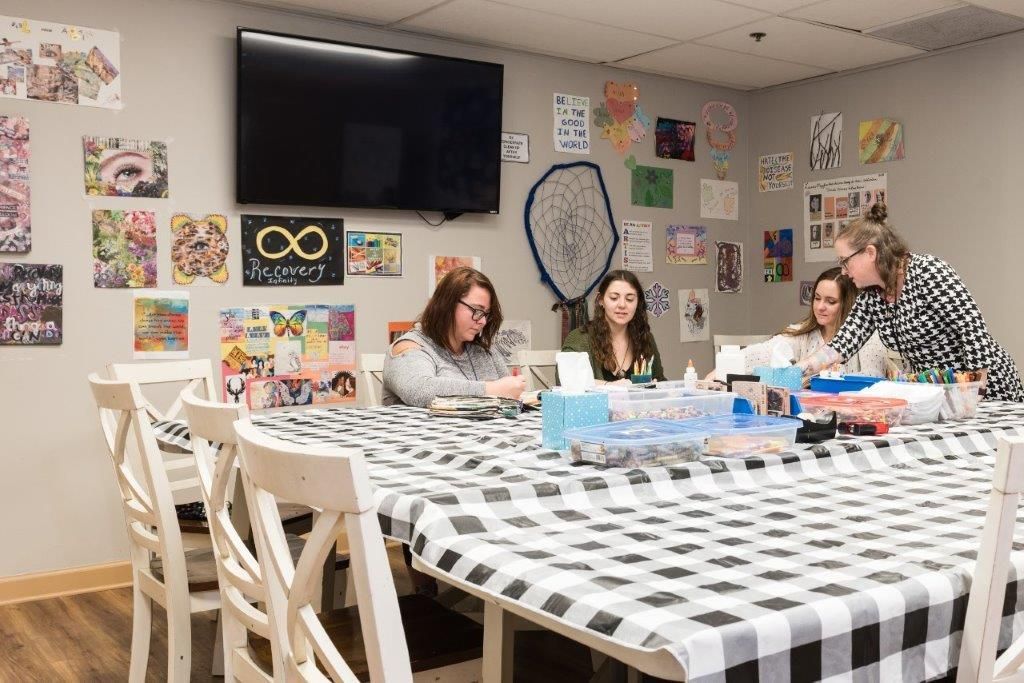 People craft at a table with a black and white checkered cloth. Artwork decorates the walls. A TV is mounted.