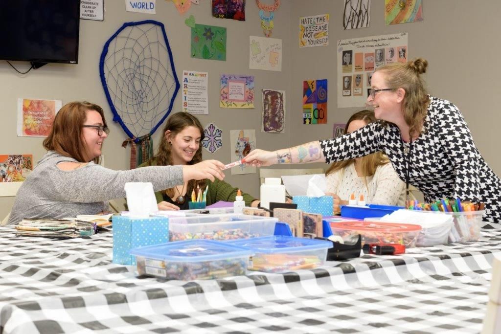 Four people crafting at a table with a black and white checkered tablecloth. They pass supplies and smile. Art decorates the walls.