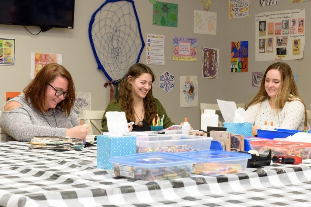 Three people craft at a table, smiling. The room has art on the walls and craft supplies.